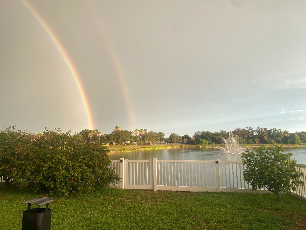 Double rainbow arching over the backyard lawn, white picket fence, and pond fountain under a grey sky