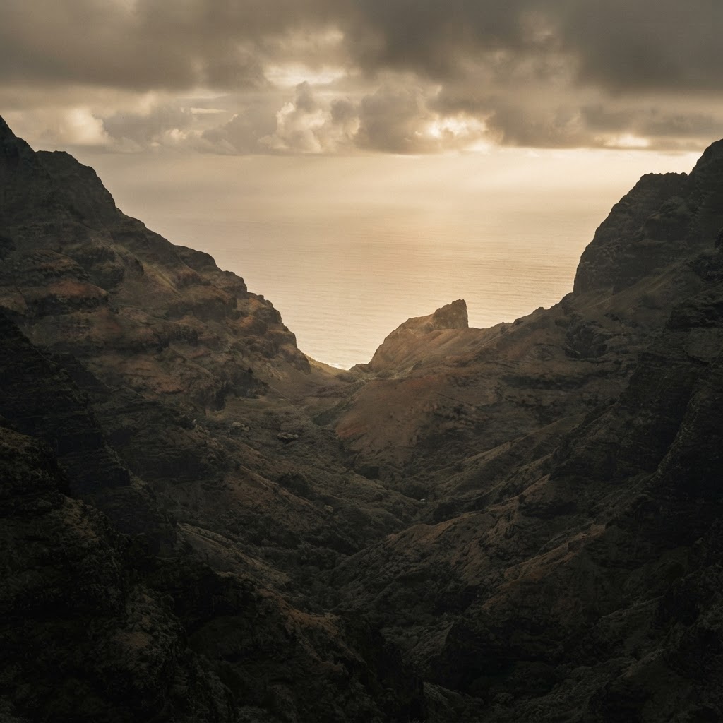 A steep, shadowed valley narrowing toward a distant opening of warm light over the ocean