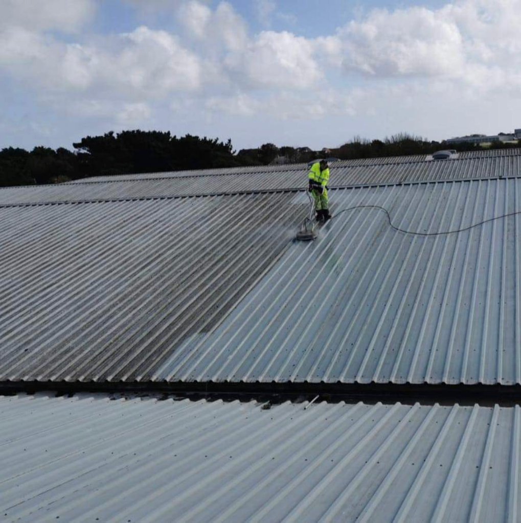 Operative pressure washing corrugated metal roof surface showing cleaned and uncleaned sections across large industrial roof.