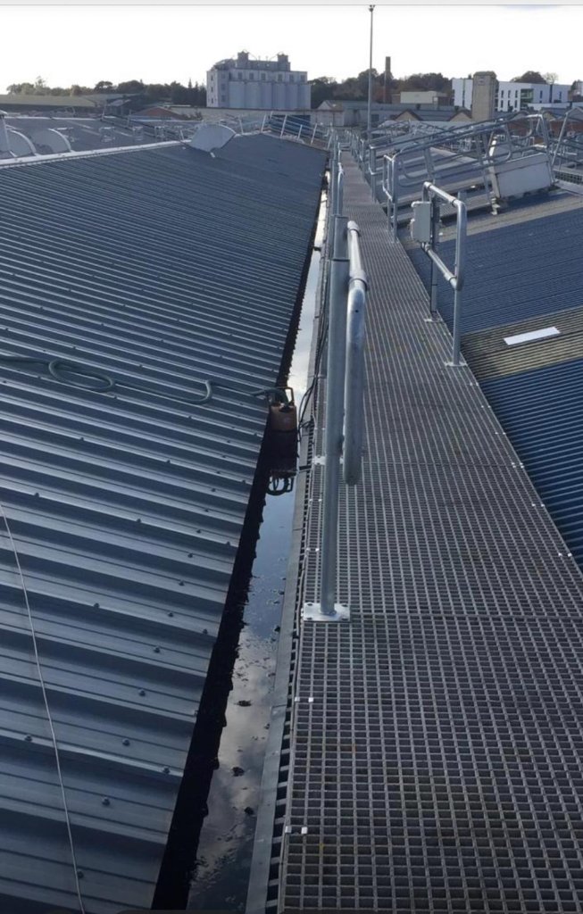 Elevated view along commercial industrial roof showing coated profiled metal surface, metal grating walkway, safety handrail and standing water in box gutter.