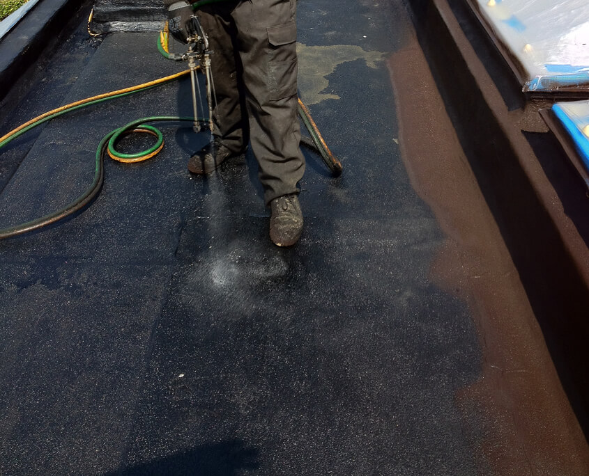 Close-up of operative applying spray waterproofing membrane to a flat roof surface, showing wet coating application adjacent to rooflight detail.
