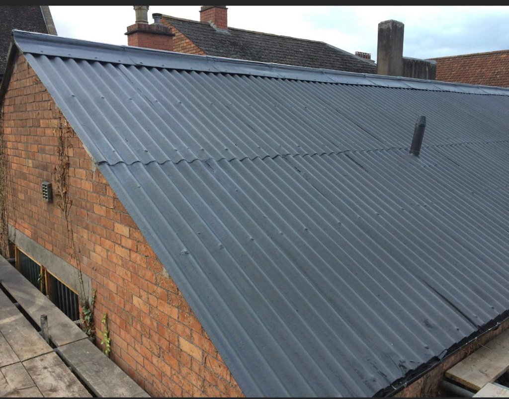 Pitched corrugated metal roof on traditional brick building coated in dark grey, showing completed refurbishment with ridge and penetration details.