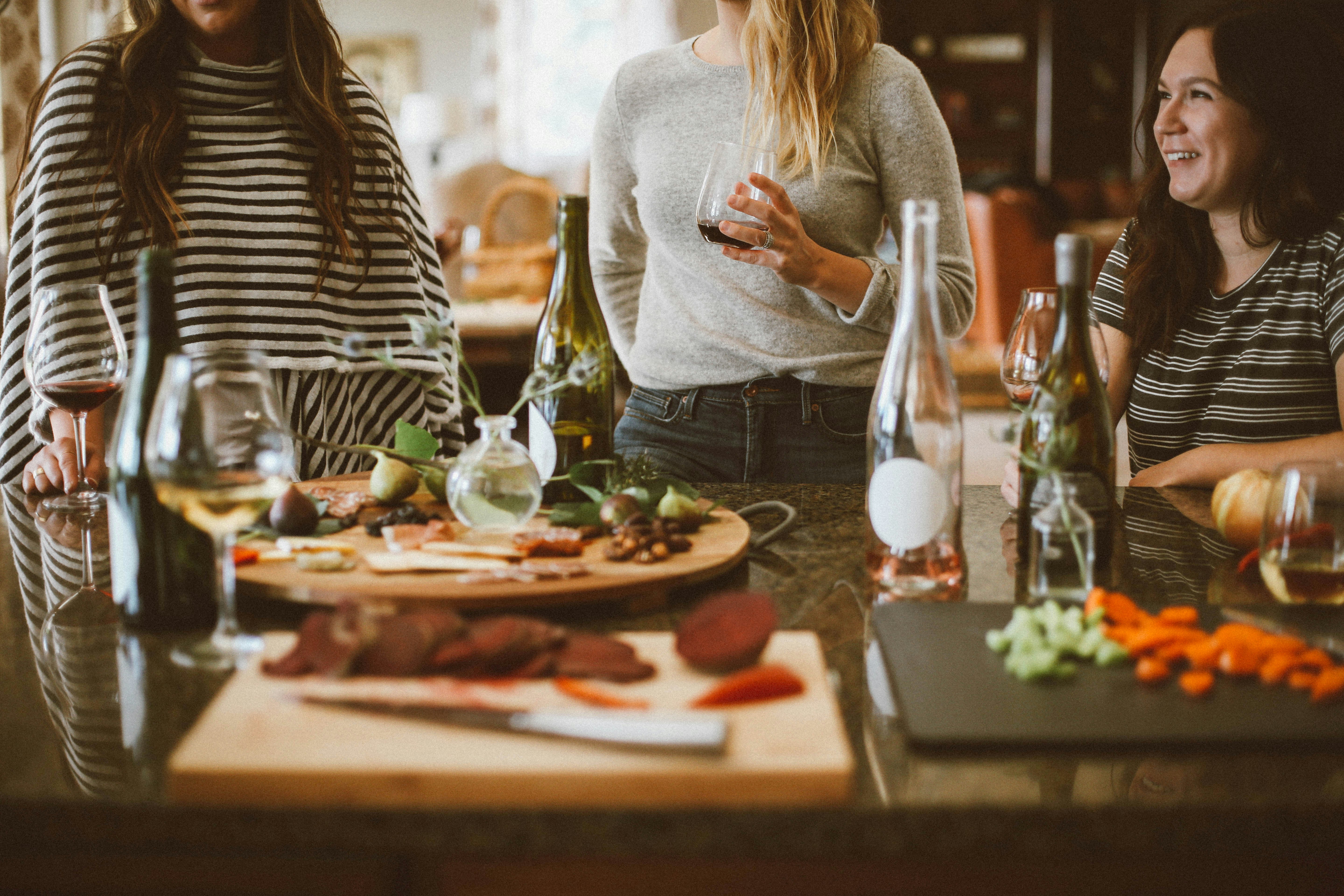 Three women gathered around a kitchen island sharing wine and food