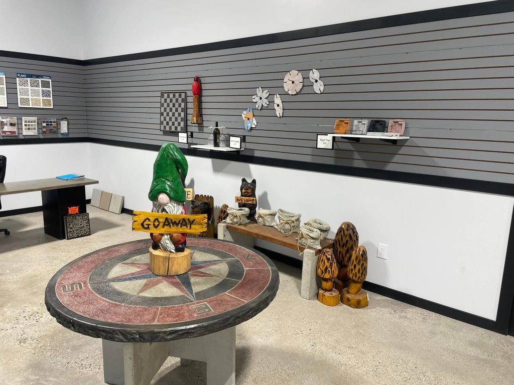 Showroom corner with a compass rose concrete table, a hand-carved wooden GO AWAY gnome, color flake samples, and concrete planters