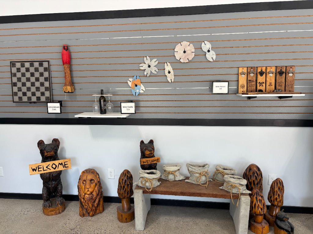 Showroom slat-wall display with a wooden checkerboard, carved cardinal, wine caddy, bottle openers, welcome bears, lion carving, and morel mushroom carvings