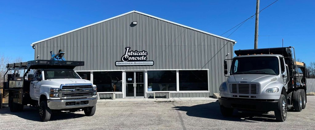 Intricate Concrete Designer Showroom exterior — grey metal-sided building with glass storefront entrance, signage reading Intricate Concrete and Designer Showroom, two work trucks parked out front