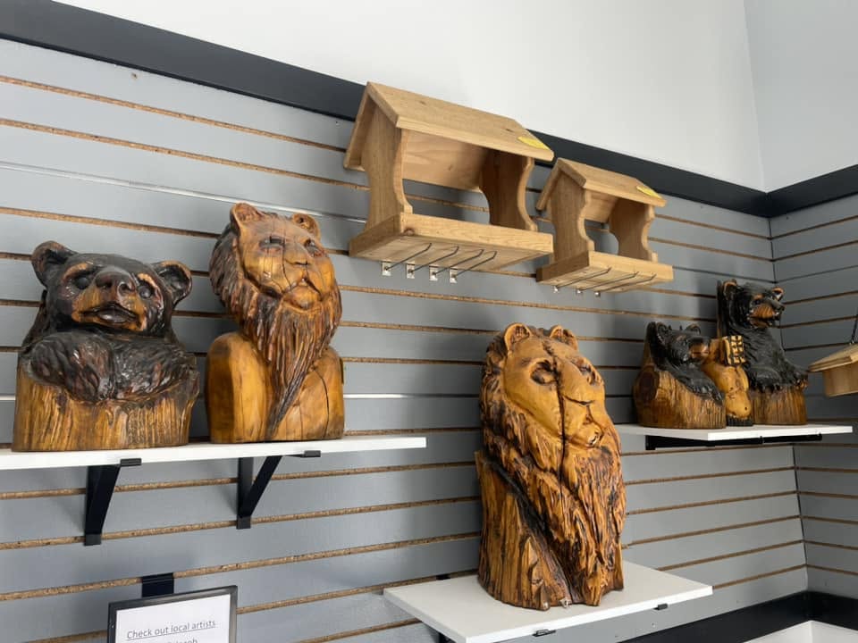 Grey slat-wall display with hand-carved wooden bear head, lion head, and lion mane sculptures alongside wooden bird feeders on floating shelves