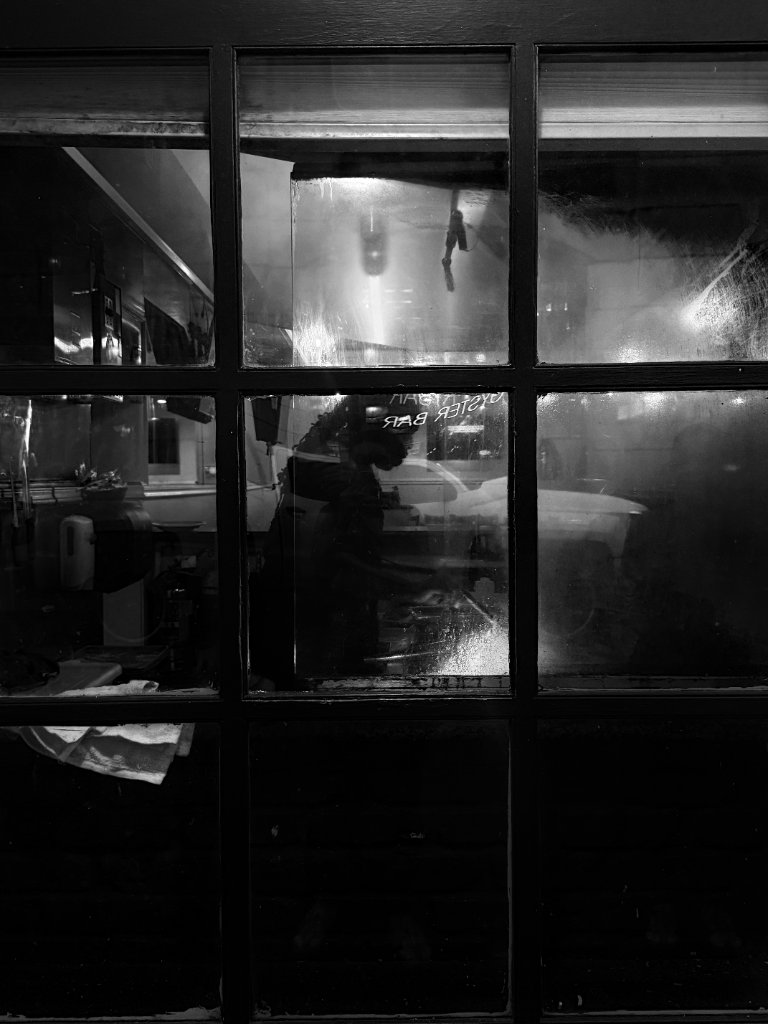 A reflection in a rain-streaked window of an oyster bar — a silhouetted figure inside, warm light glowing through the glass