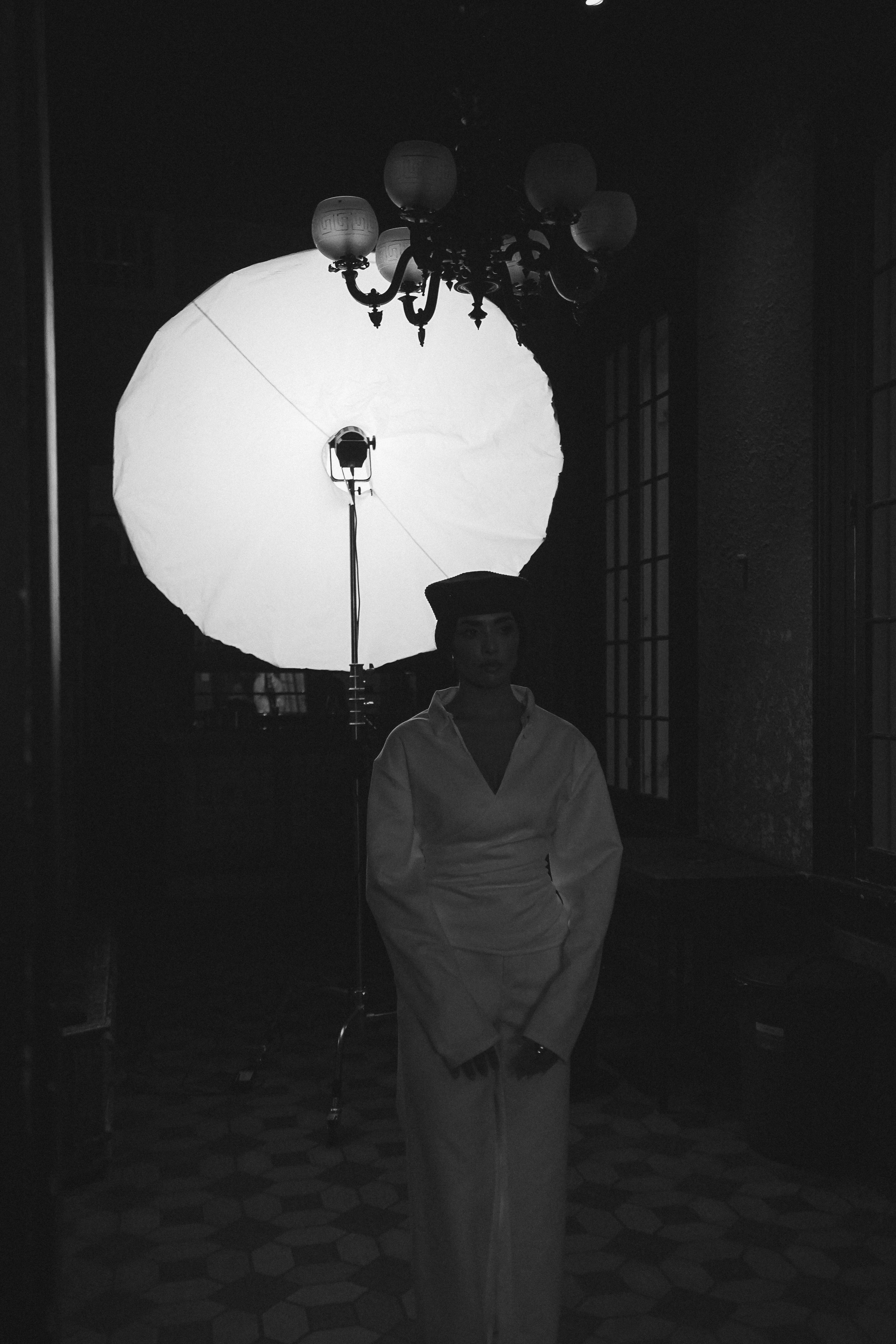 A woman in a white outfit and dark hat standing before a large circular softbox with a chandelier above, black and white portrait