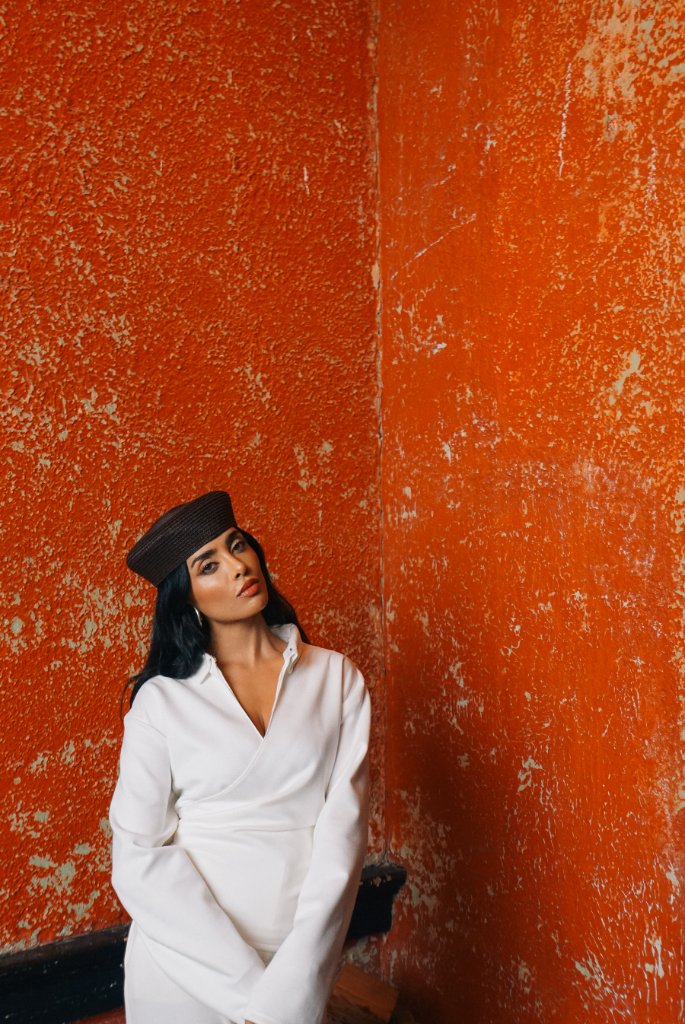 A woman with dark hair in a brown pillbox hat and white collared shirt with silver hoop earrings against a heavily textured distressed orange wall with a vertical corner