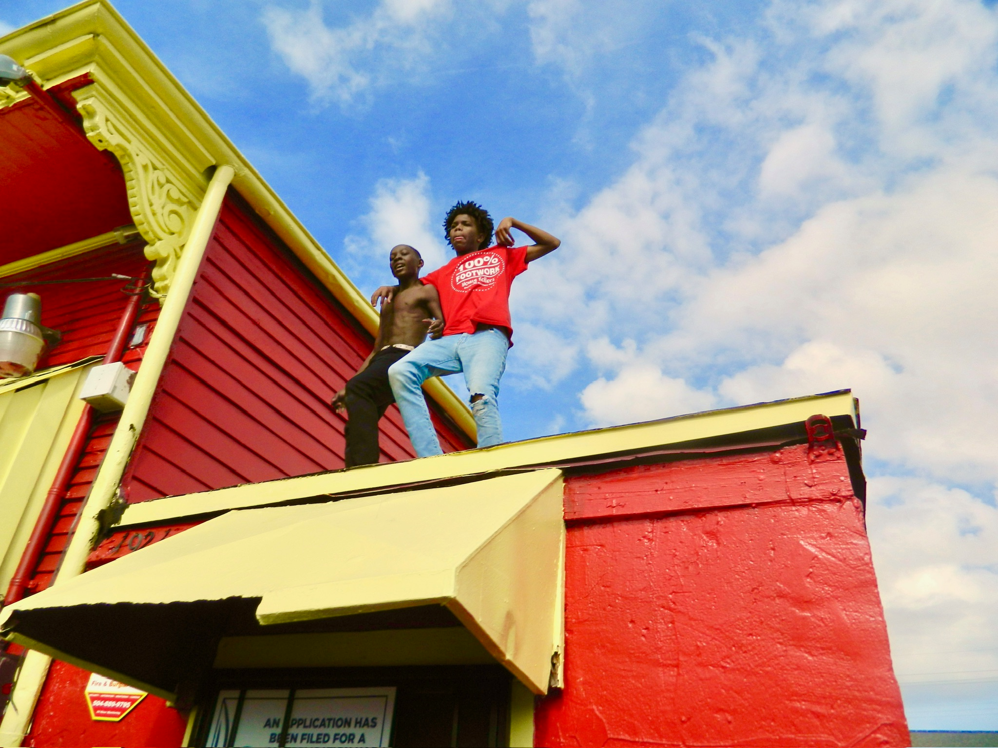 Two young boys standing on the roof of a bright red building with yellow trim, one shirtless and one in a 100% FOOTWORK t-shirt, clear blue sky behind