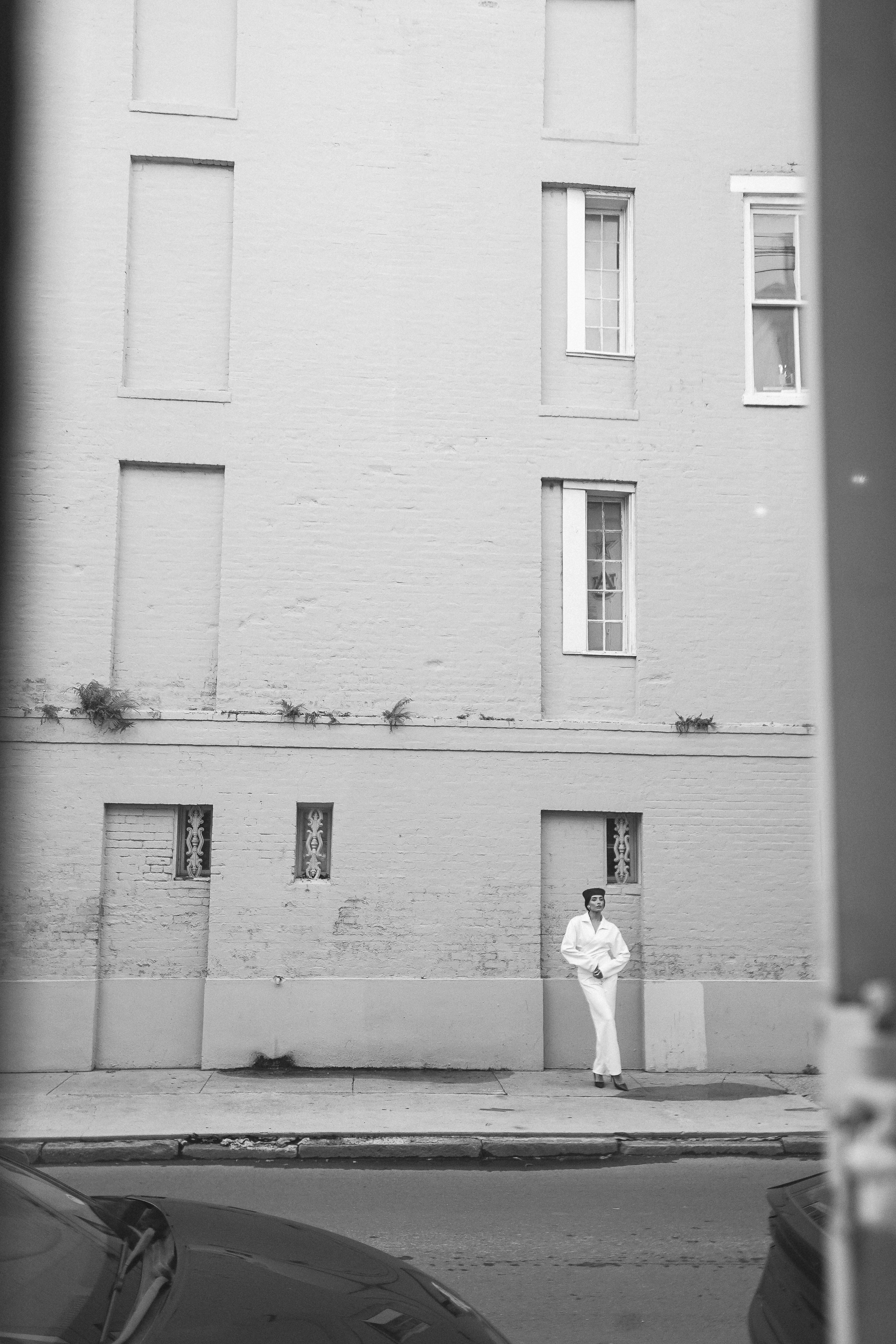 A woman in a white wrap shirt and trousers with a dark pillbox hat standing against a textured brick building facade, black and white full-body portrait