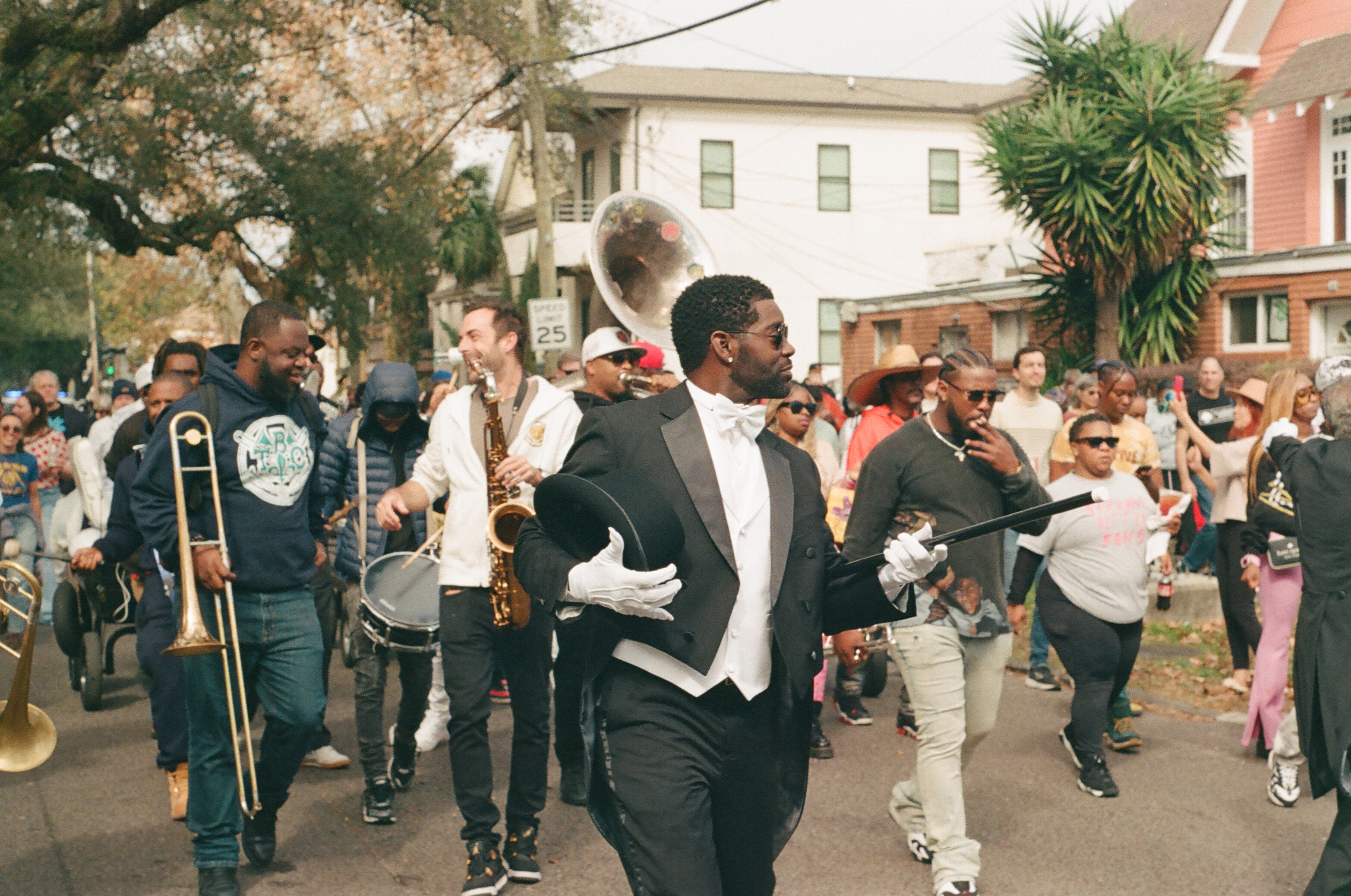 A street parade band in a residential neighborhood — a man in a black tuxedo tailcoat with white bow tie and gloves holding a hat and cane, musicians with trombone and saxophone, palm tree and beige houses in the background