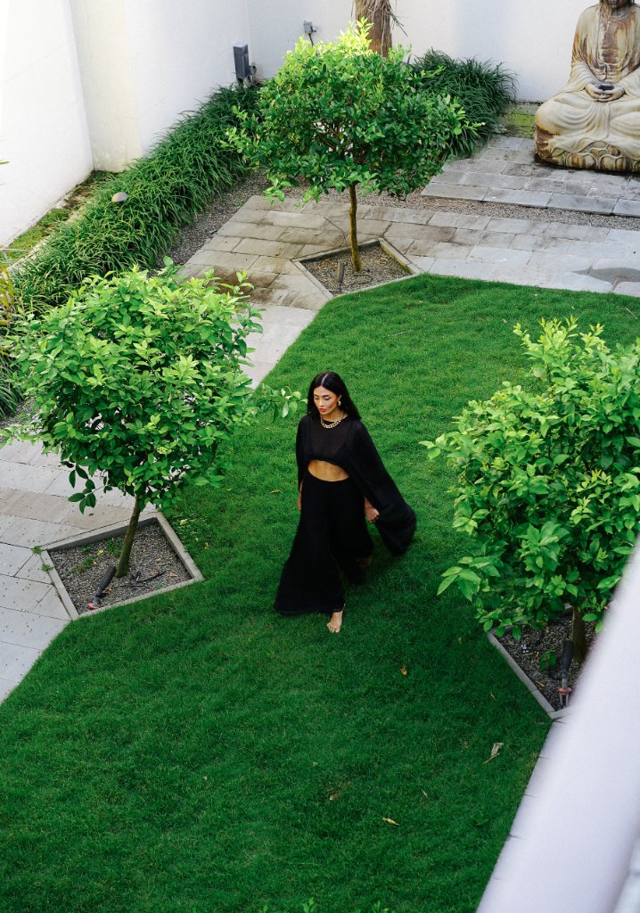 A woman in a black crop top and wide-leg trousers walking barefoot across a green courtyard lawn between two small trees, stone-paved walkway, seated Buddha sculpture in background