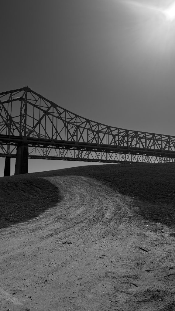 A dirt levee path leads toward a steel truss bridge stretching across the sky, harsh midday sun, New Orleans