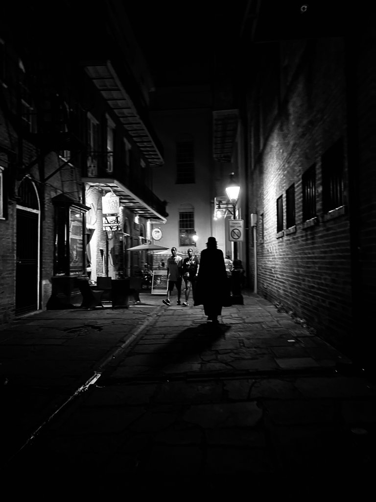 A silhouetted figure walks down a dark New Orleans alley at night, lit by a single street lamp