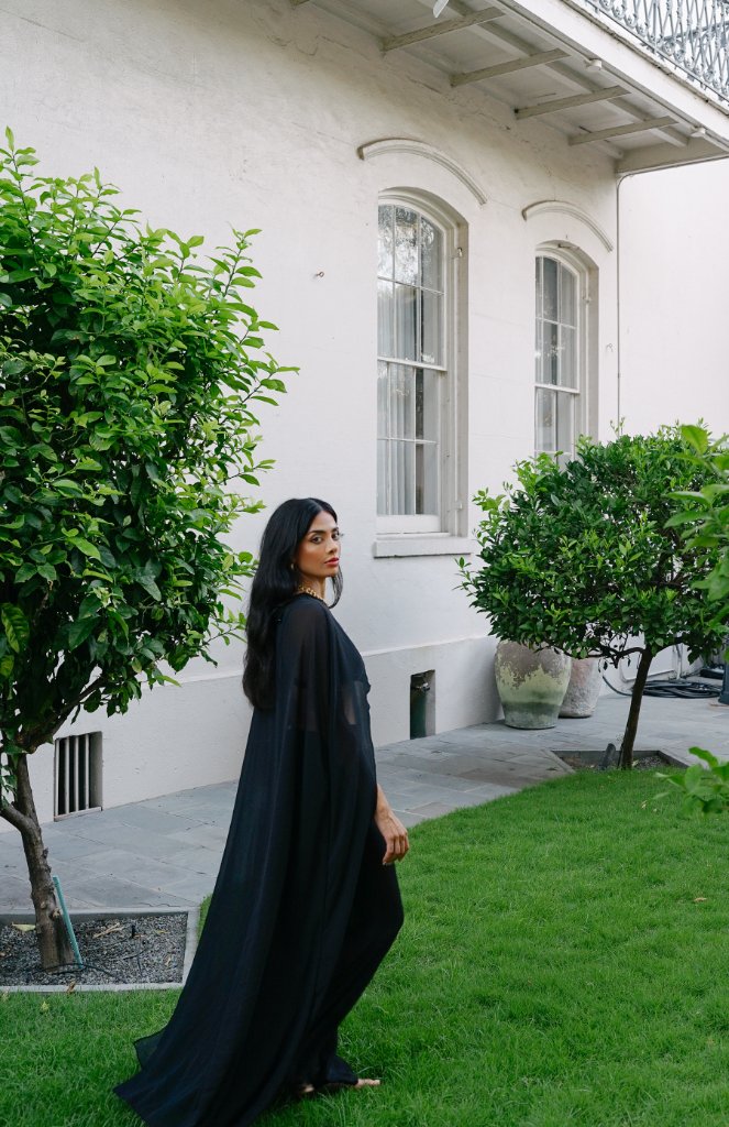 A woman in a flowing semi-transparent black cape over a black top and pants in profile, standing on a stone path beside grass, white building wall with arched windows behind, green trees
