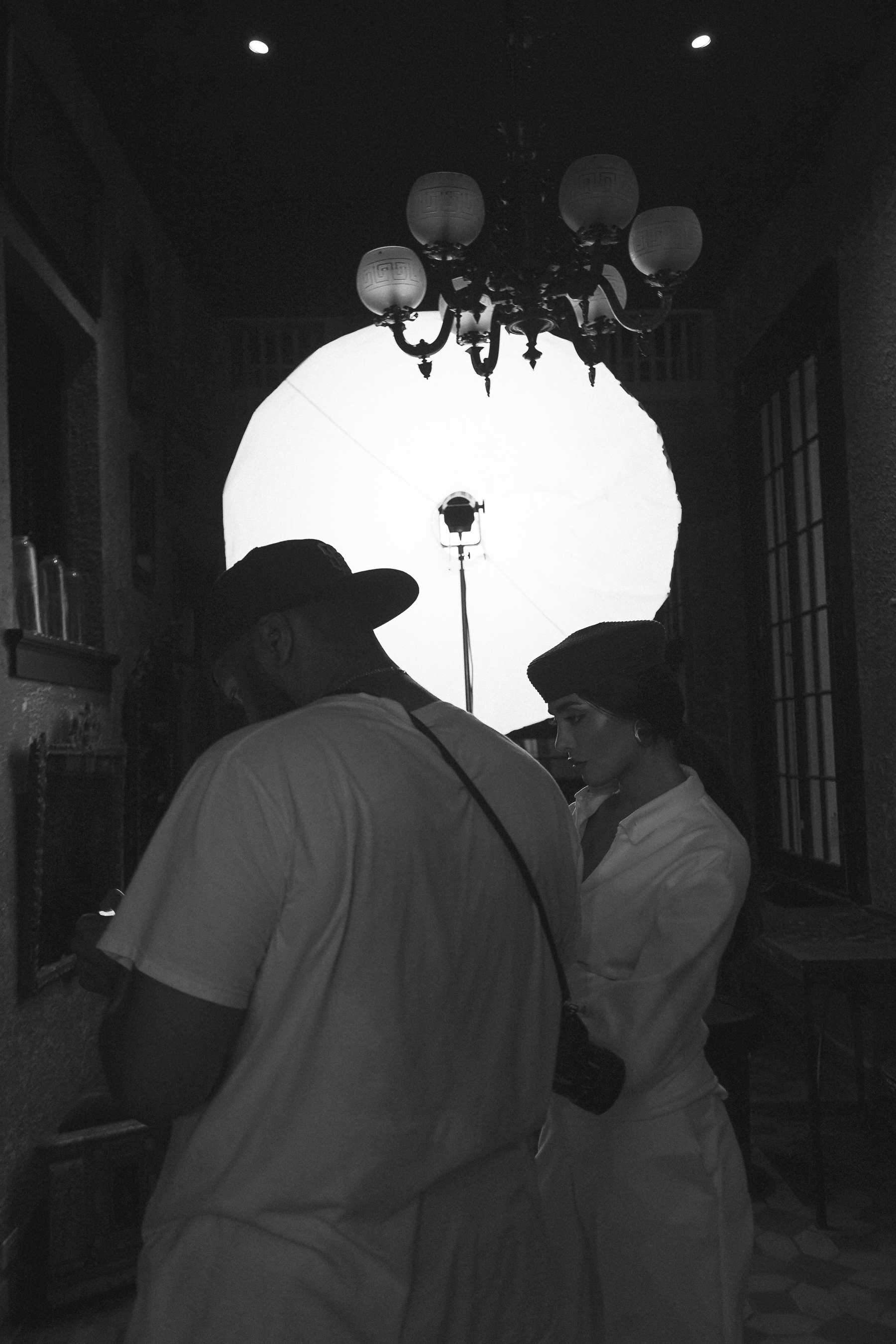 A photographer in a cap looking down at his camera beside a woman in profile in a dark hat and white jacket, a glowing softbox and chandelier behind, black and white