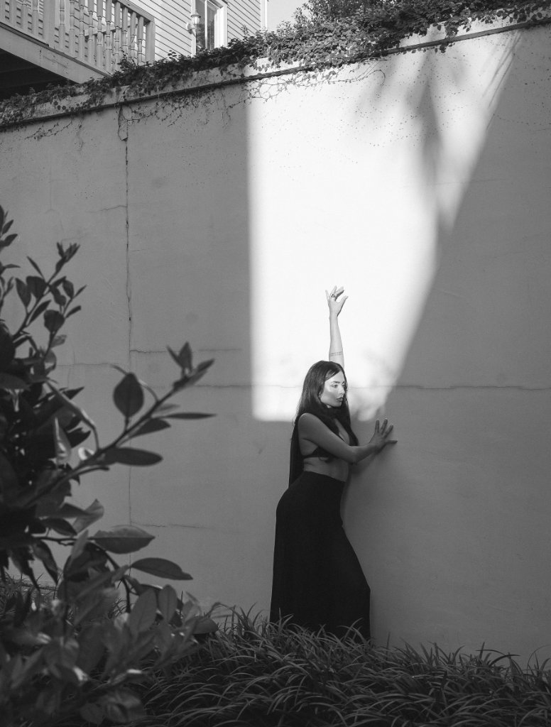 A woman in a dark sleeveless top and flowing skirt in profile, arm raised toward a wall hit by a vertical beam of light, foliage in the foreground, high-contrast black and white