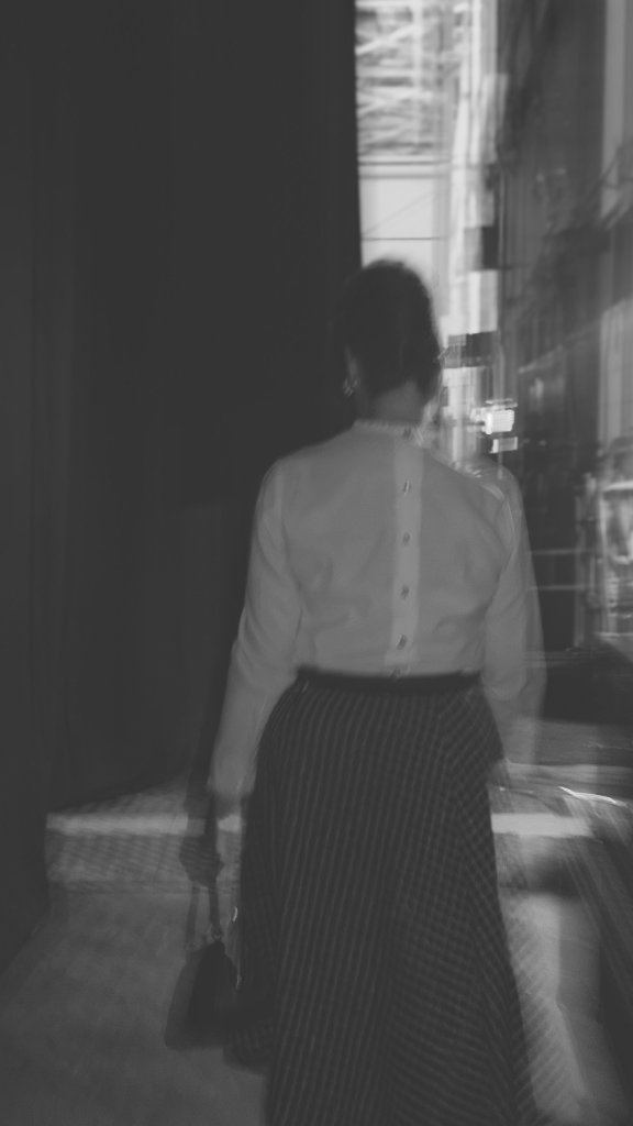 A blurred black and white figure of a woman in a white blouse and striped skirt walking through a dark corridor