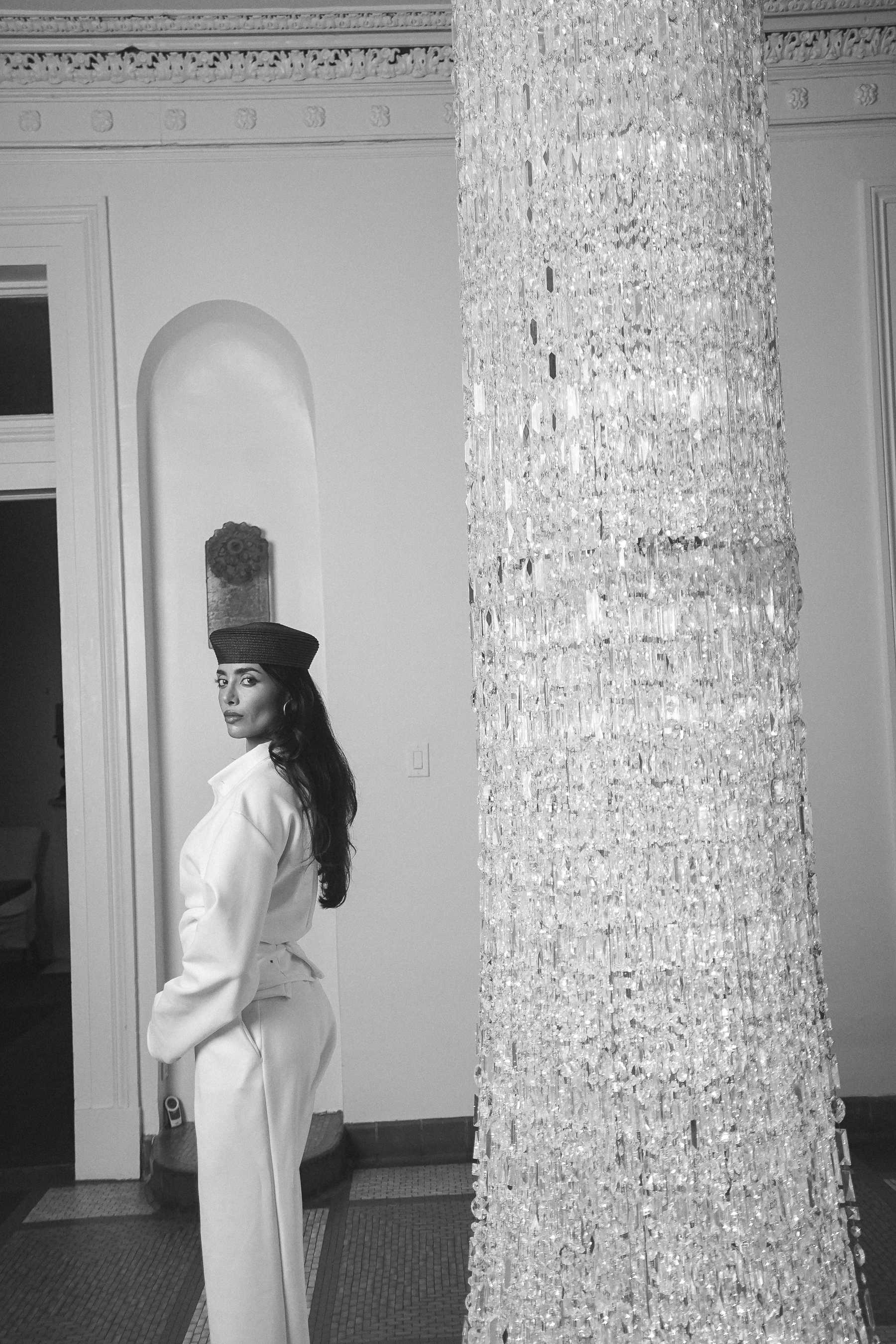 A person in a white wrap jacket and trousers with a dark pillbox hat standing in profile beside a tall crystal column, black and white portrait