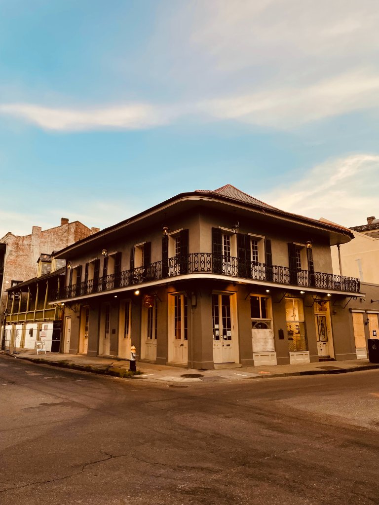 A golden corner building with ornate iron balconies and tall windows, lit by golden hour sunlight, New Orleans French Quarter architecture