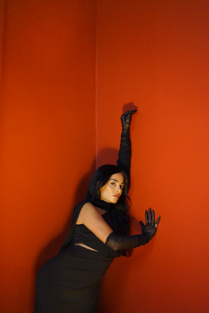 A woman in a black dress and gloves standing against a corner of bright terracotta-red walls, right arm raised high, left hand pressed against the wall