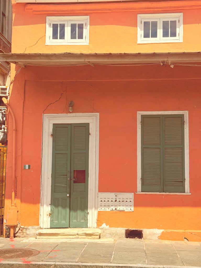 A vivid matte orange two-story building facade with sage green shutters and door, cream window frames, and metal mailboxes mounted on the wall