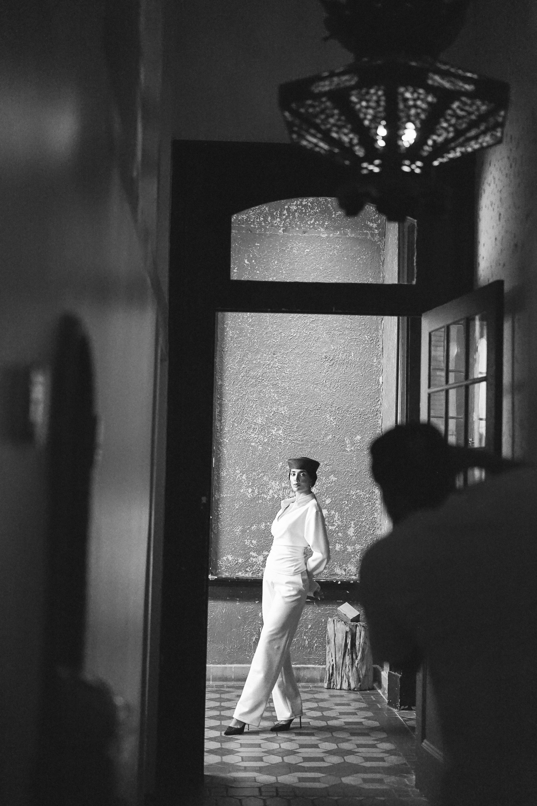 A woman in a white outfit and dark pillbox hat framed by a doorway in a textured room, with a photographer silhouette in the foreground, black and white