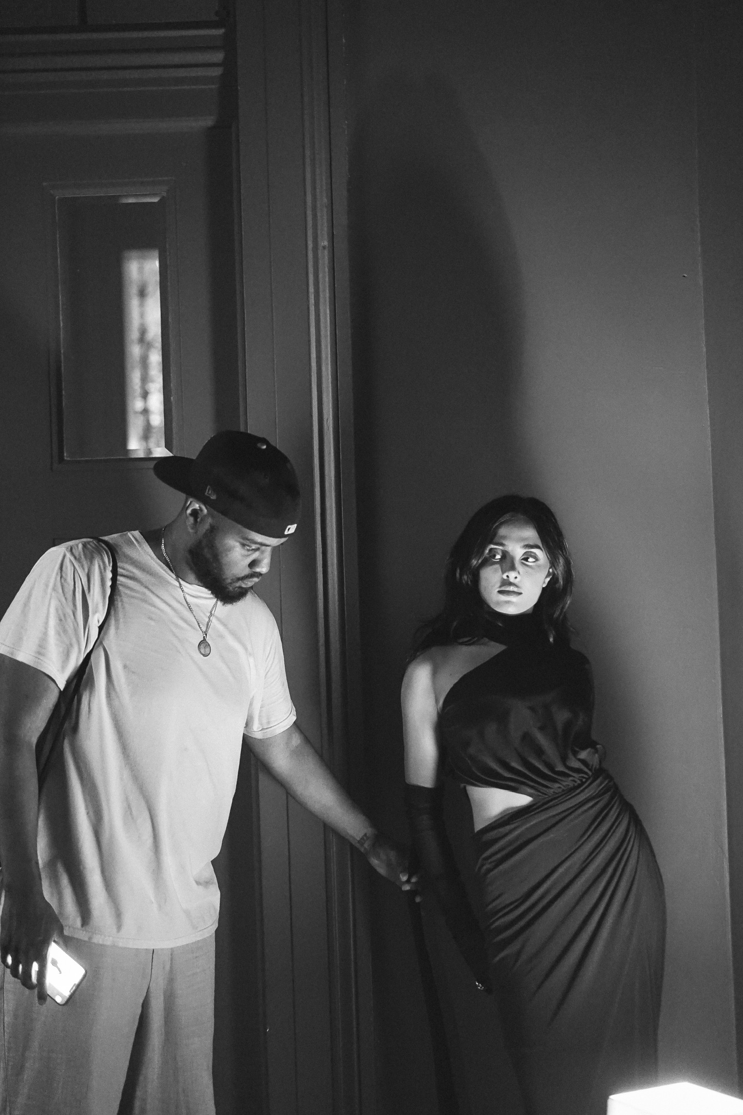 A photographer in a cap reaching toward a model in a dark one-shouldered dress and gloves against a wall, black and white behind the scenes