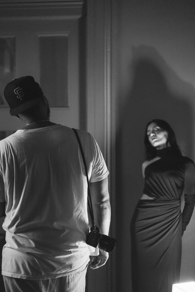 A behind-the-scenes photoshoot scene — a photographer in a cap and T-shirt on the left, a model in a draped dress standing against a wall on the right, high-contrast cinematic black and white