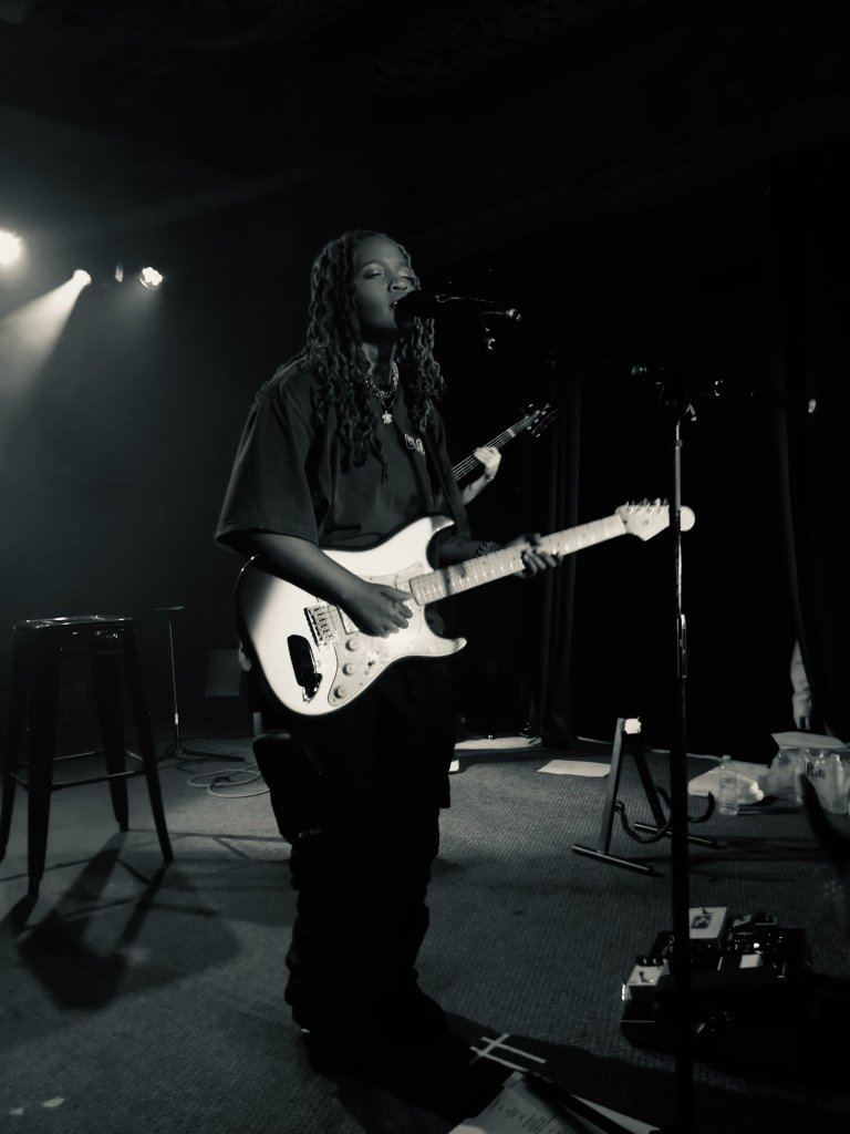 A guitarist with long braided hair playing an electric guitar and singing on a dark concert stage, dramatic stage light with lens flare, black and white