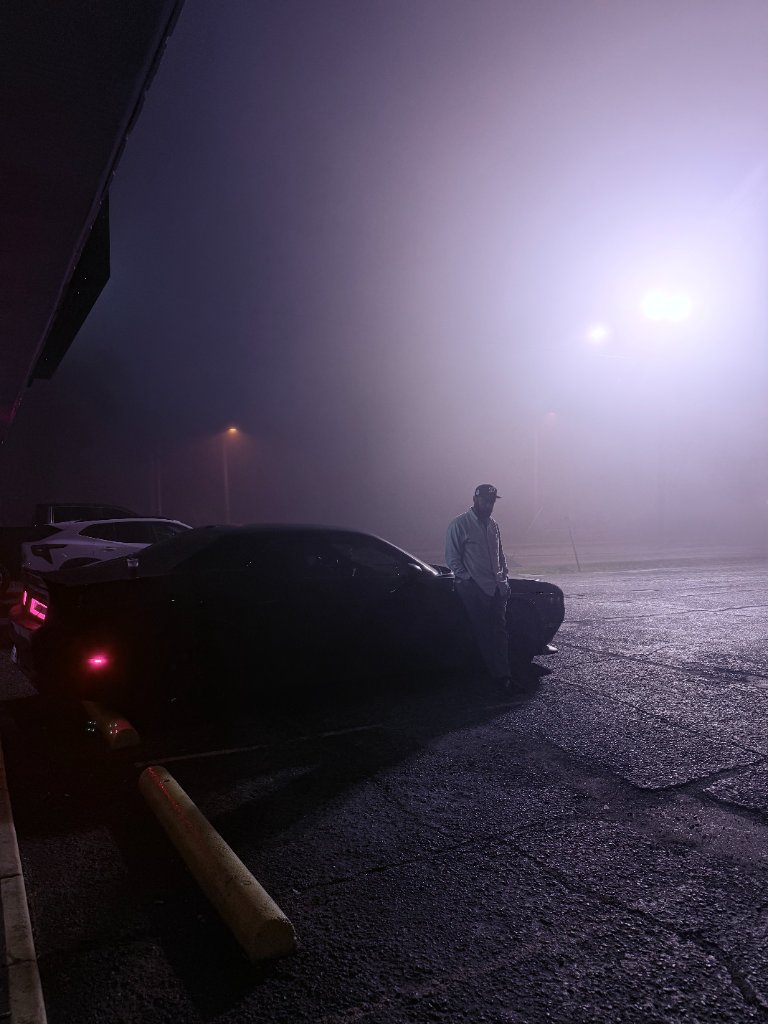 A moody foggy night parking lot scene with a figure standing by a car under purple and white atmospheric lighting, minimal visibility