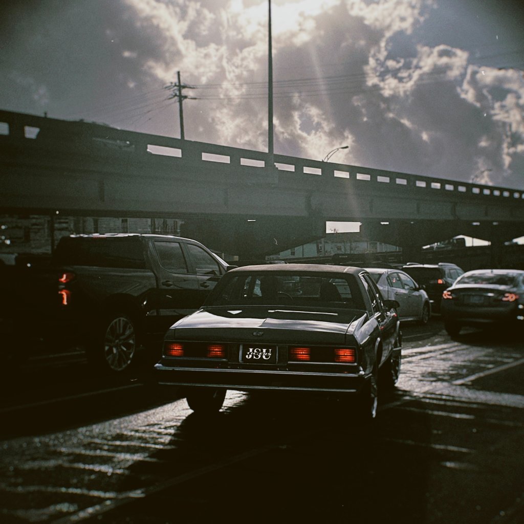 A classic black car waits at a traffic light beneath an overpass, dramatic backlit sky