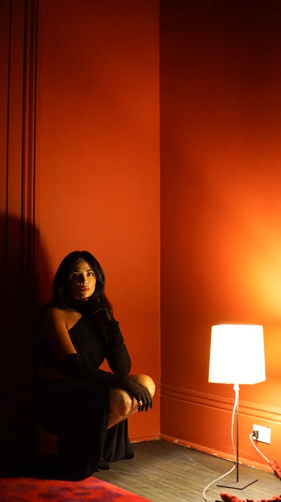 A woman in a black one-shoulder dress and sheer gloves crouching low near a corner of deep orange-red walls lit by a floor lamp