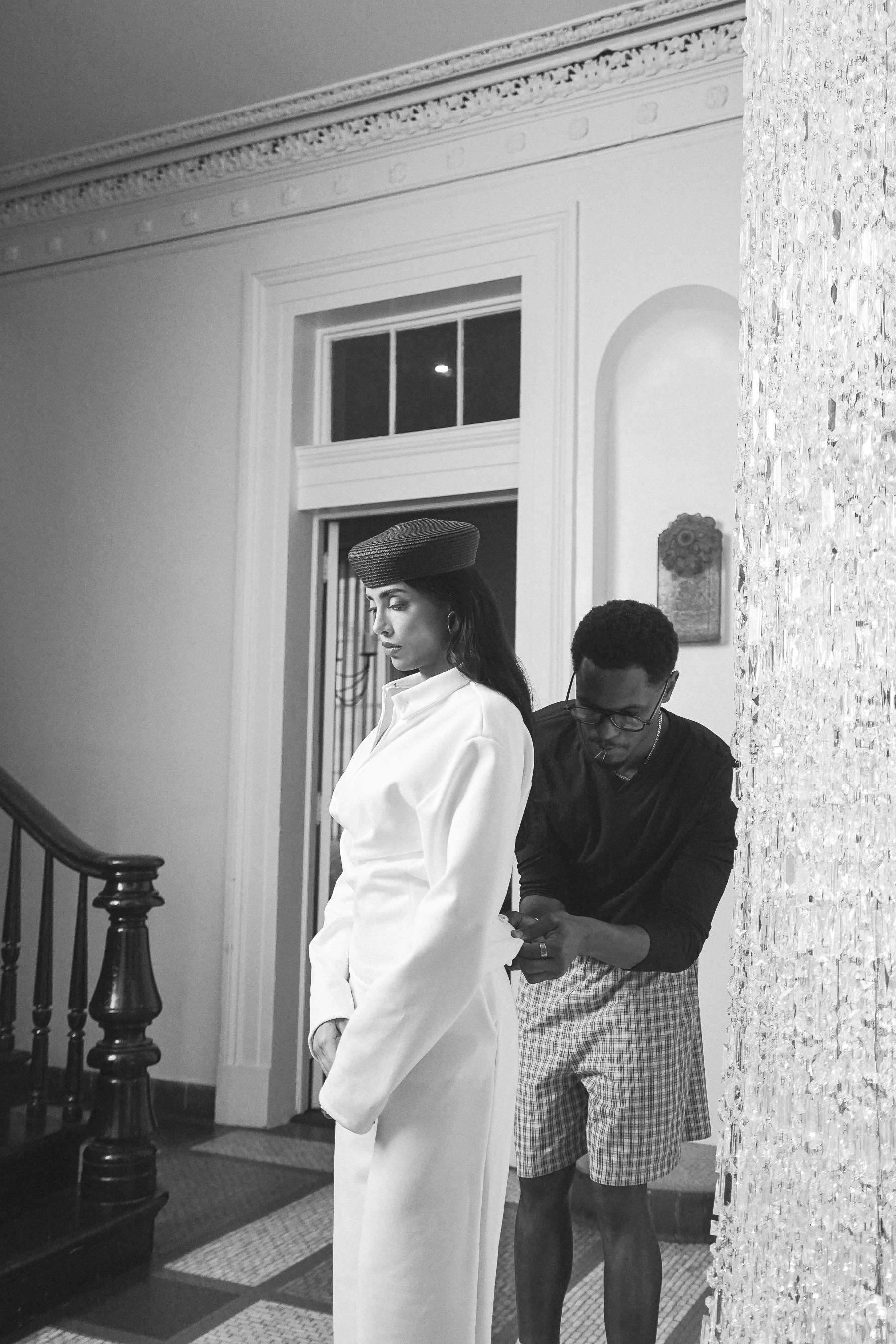 A woman in a white outfit and dark pillbox hat standing in profile in a hallway while a man adjusts her top, a glowing crystal chandelier beside them, black and white