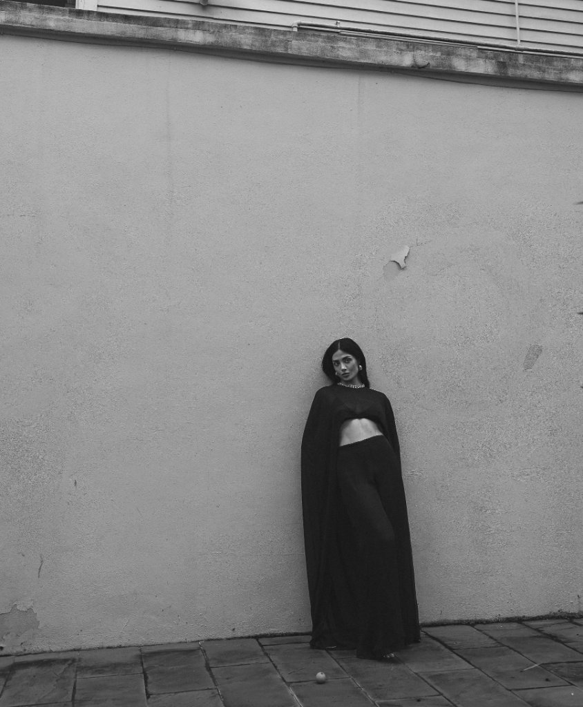 A woman in a dark crop top and flowing trousers leaning against a textured wall, full-body black and white portrait
