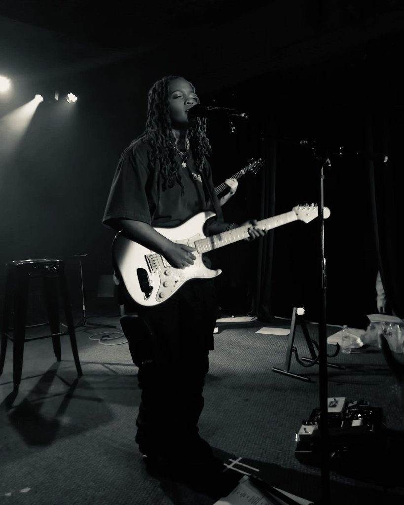 A singer plays electric guitar on stage under dramatic spotlighting, singing into a microphone, black and white concert performance