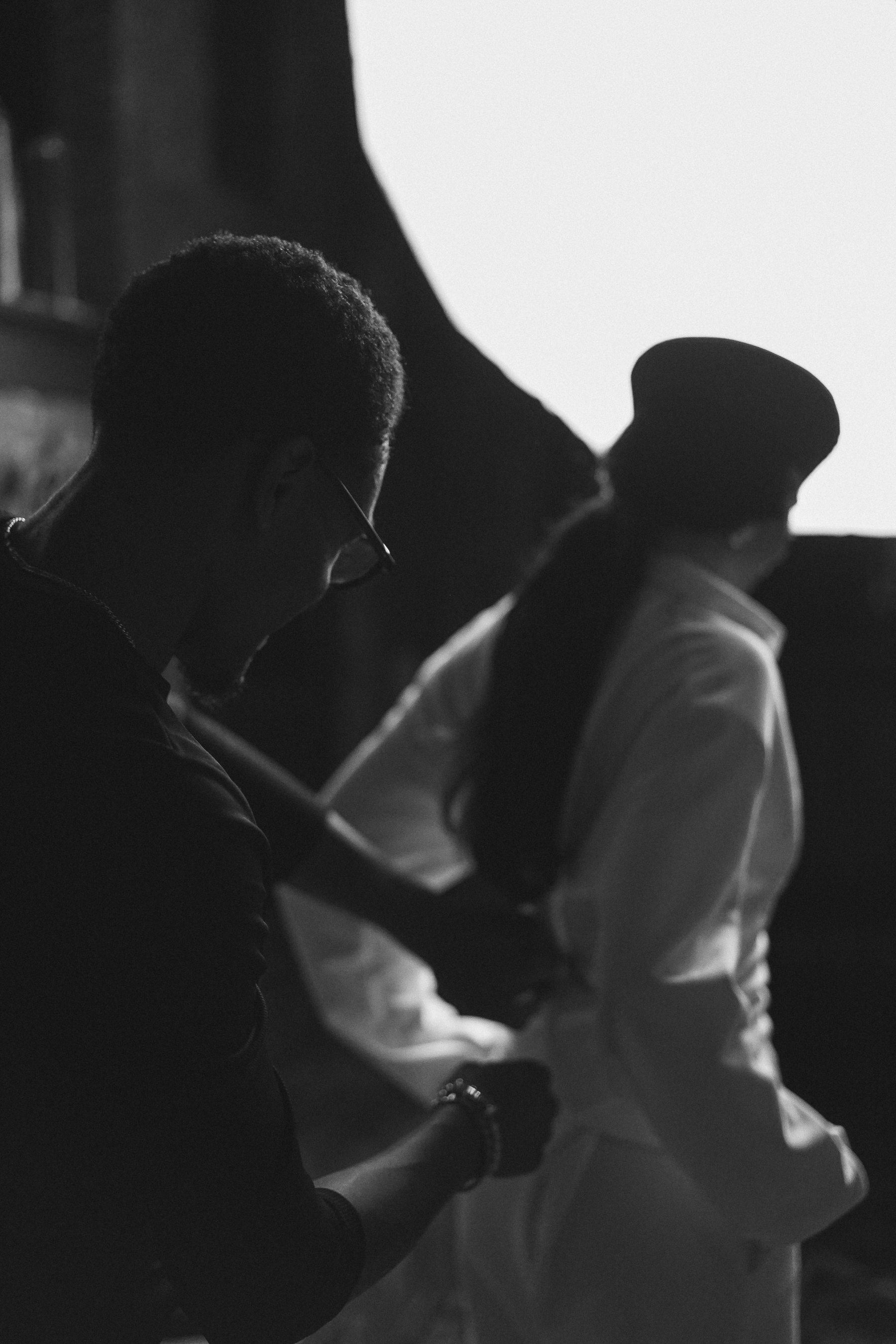 Two figures from behind, a person adjusting the waist of a woman in a white garment and dark hat, backlit by a bright circular light, black and white