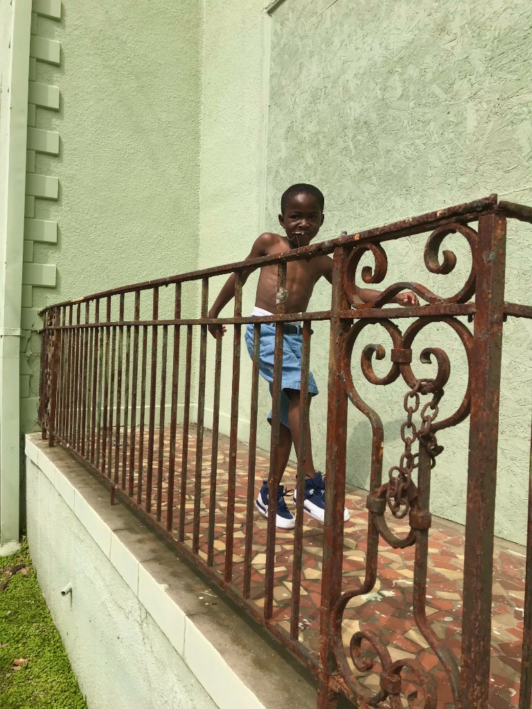 A young shirtless boy in denim shorts leaning against a rusted ornate metal railing, sage green wall and mosaic tile floor, soft daylight