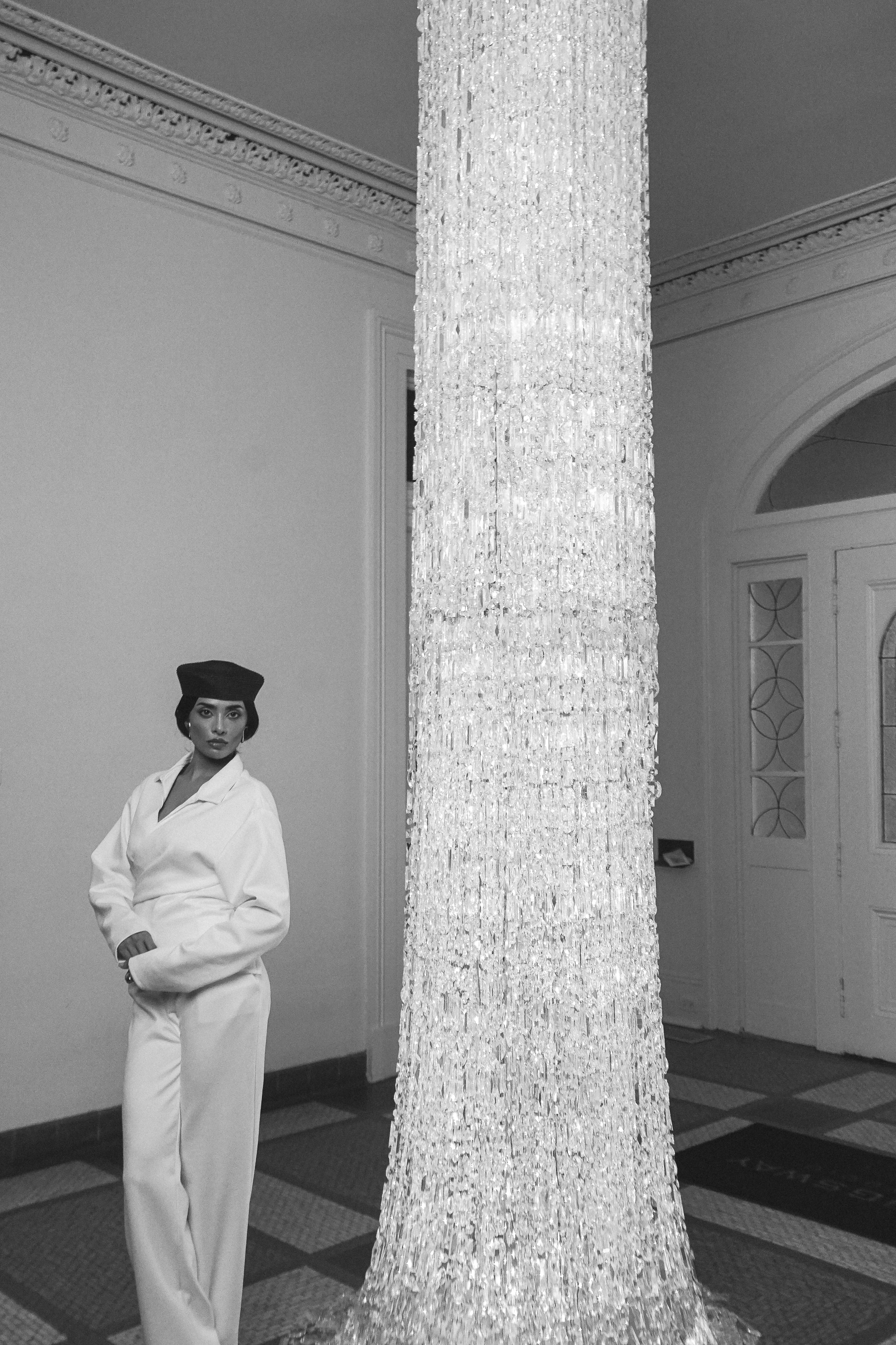 A woman in a white pantsuit and dark cap standing beside a tall ornate chandelier in an arched doorway, black and white portrait