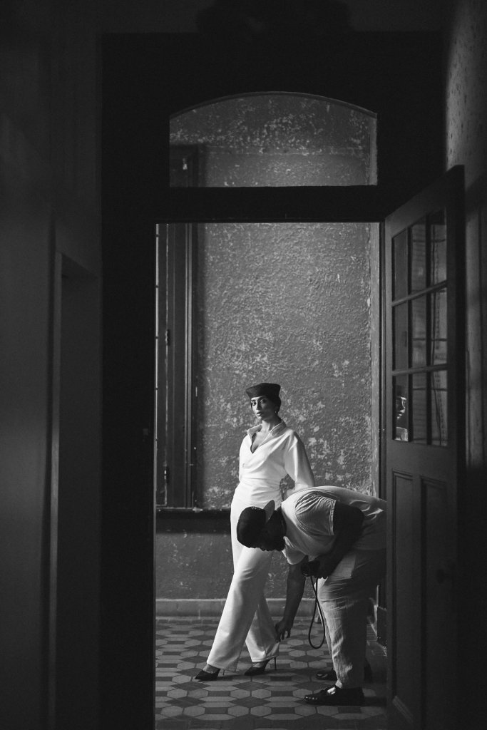 A person in a white collared shirt and dark pillbox hat standing in a doorway corridor, dramatic black and white portrait