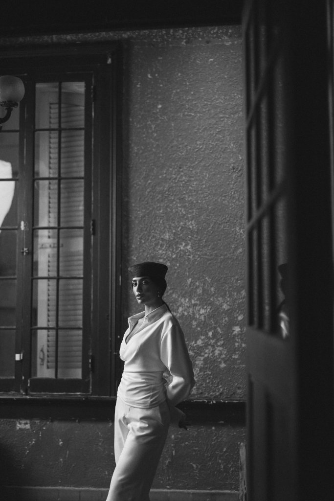 A woman in a structured white top and dark pillbox hat standing in an interior alcove, cinematic black and white portrait