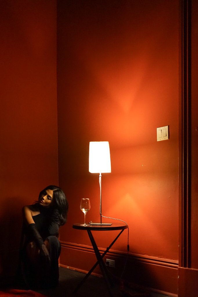 A woman in a black outfit and gloves crouching low near a corner of deep burnt-orange walls lit by a lamp on a small side table with a wine glass