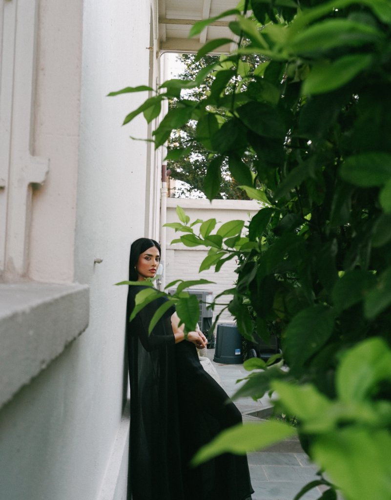 A woman with long dark hair in a black outfit standing against a cream-toned exterior wall, large out-of-focus green bush to the right, soft daylight