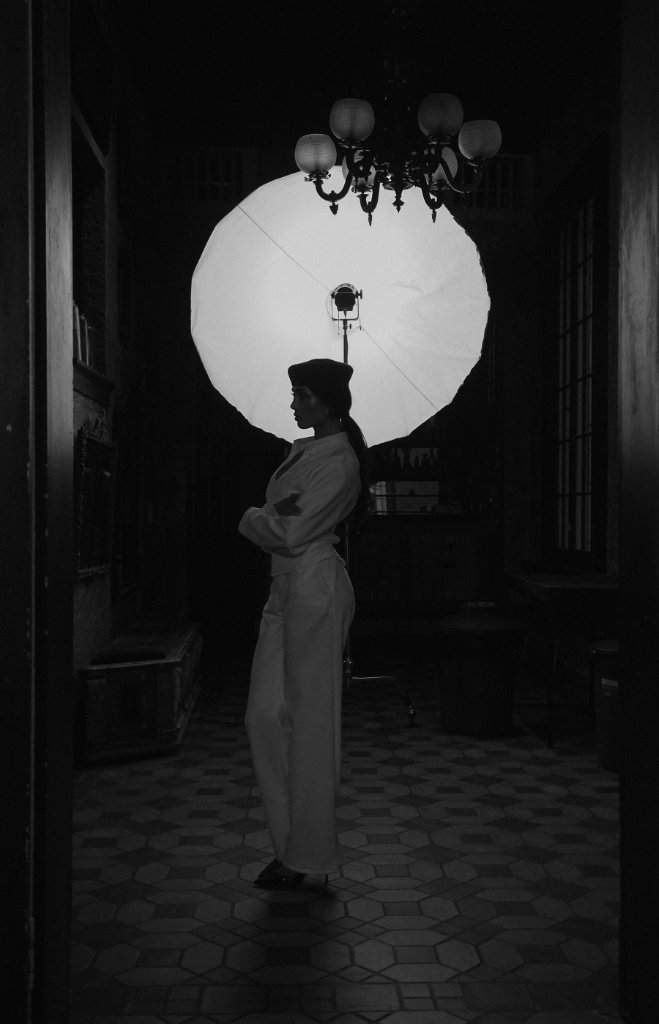 A woman in a beret and collared shirt in profile, arms crossed, backlit by a circular softbox in a dim hallway with a chandelier above, high-contrast black and white