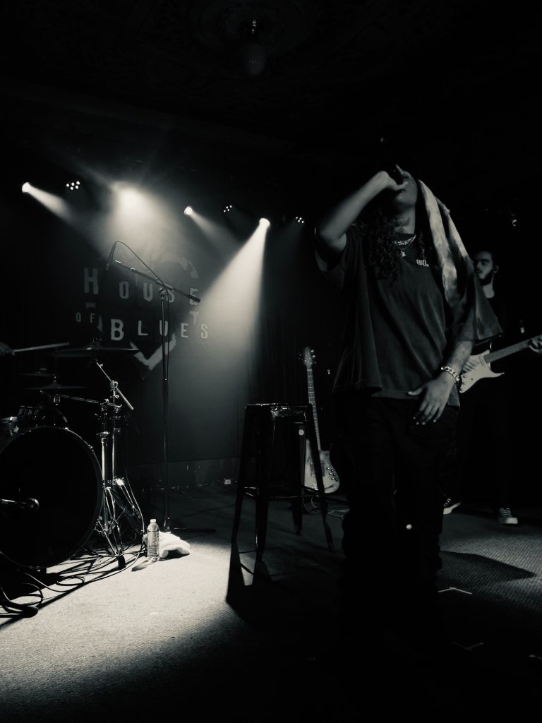 A performer with long curly hair singing into a microphone at House of Blues, dramatic spotlighting, black and white concert photography