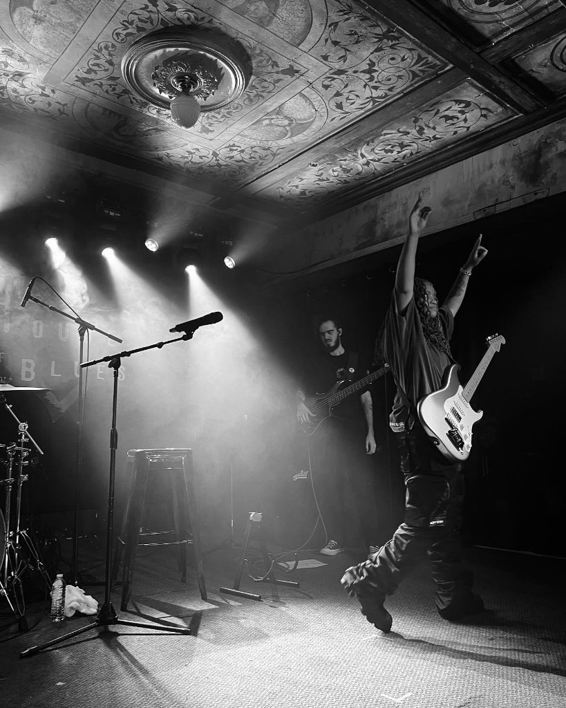 A guitarist raises his arm on a smoky stage beneath an ornate ceiling, dramatic stage lighting, black and white concert photography