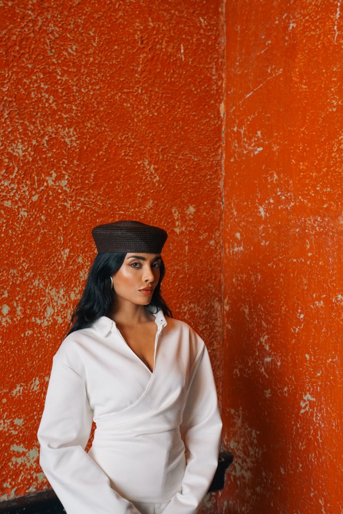 A woman with dark hair in a dark brown pillbox hat and white wrap-style top against a textured vibrant orange wall, soft directional lighting