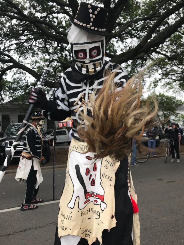 A street performer in a skeleton costume with a top hat and staff, black and white bone detailing, handwritten apron reading HELL IS HERE